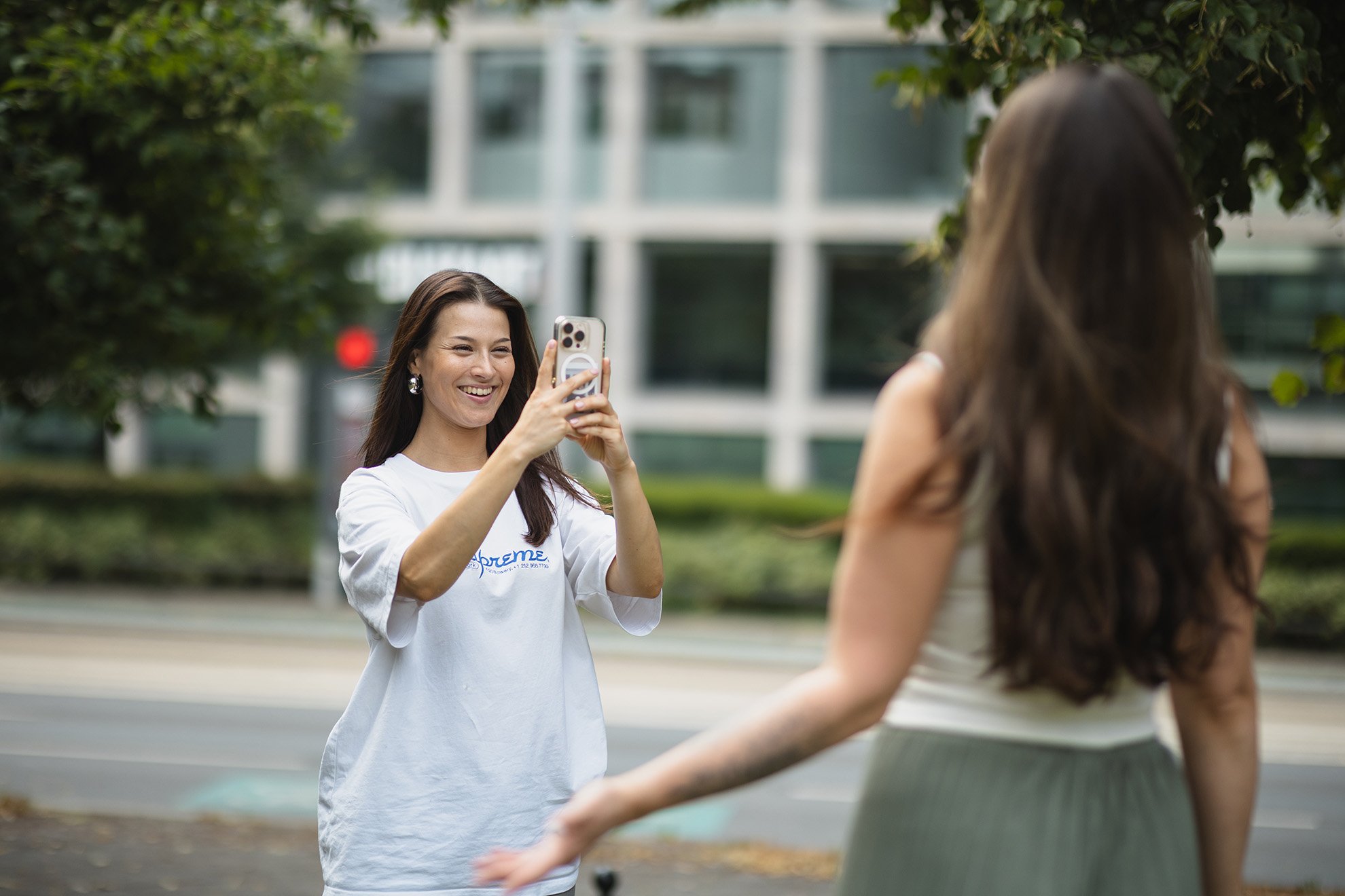 Eine junge Frau mit langen dunklen Haaren und weißem T-Shirt steht draußen und fotografiert lachend eine andere Frau mit dem Smartphone. Im Hintergrund sind Bäume und ein modernes Gebäude mit großen Fenstern zu sehen.